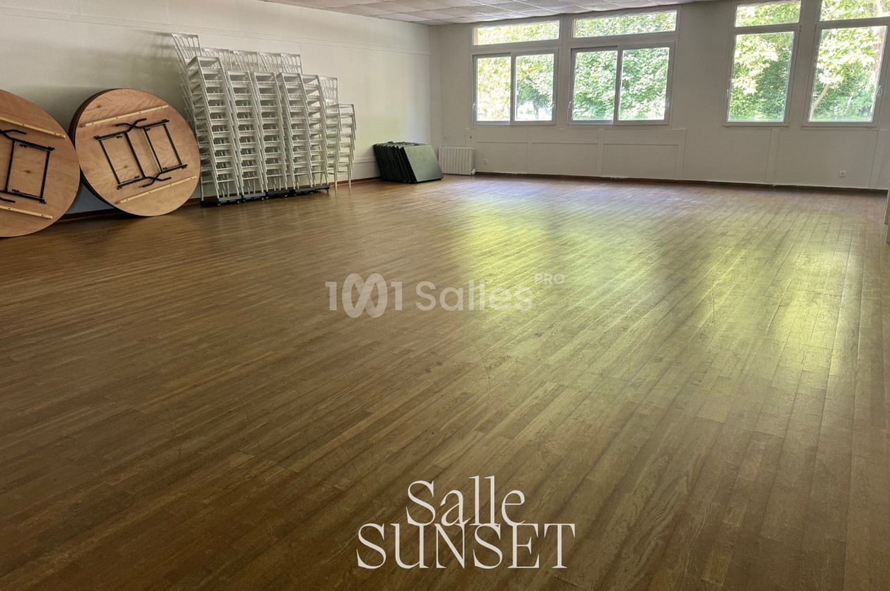 Salle vide avec parquet en bois clair, tables rondes empilées, chaises pliantes et grandes fenêtres laissant entrer la…