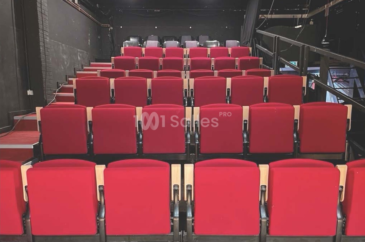 Salle de spectacle avec des rangées de sièges rouges vides et une scène visible en arrière-plan.