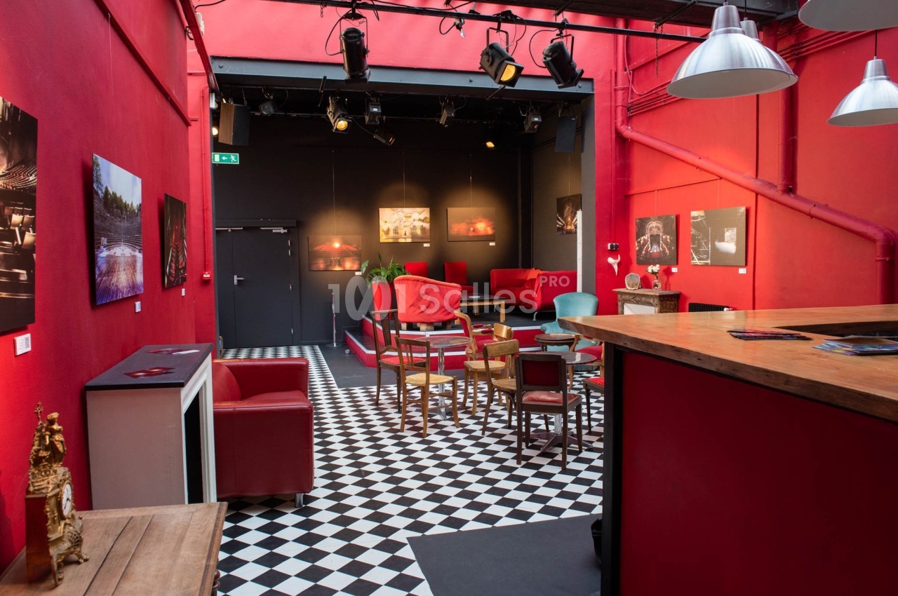 Salle avec murs rouges, sol en damier noir et blanc, mobilier varié et scène en fond éclairée par des projecteurs.