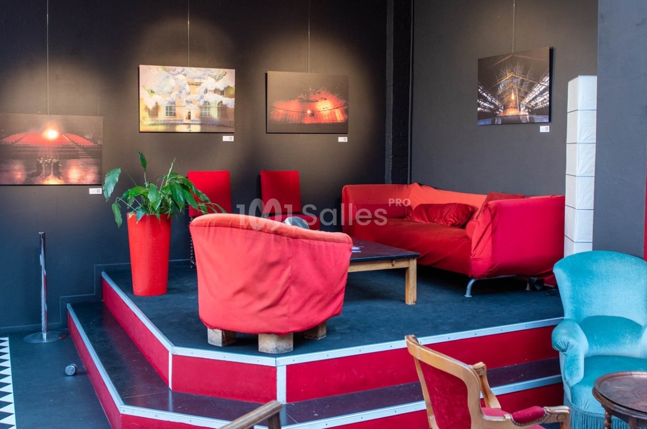 Espace salon avec canapés et fauteuils rouges, table basse en bois, plantes et photos encadrées sur un mur sombre.