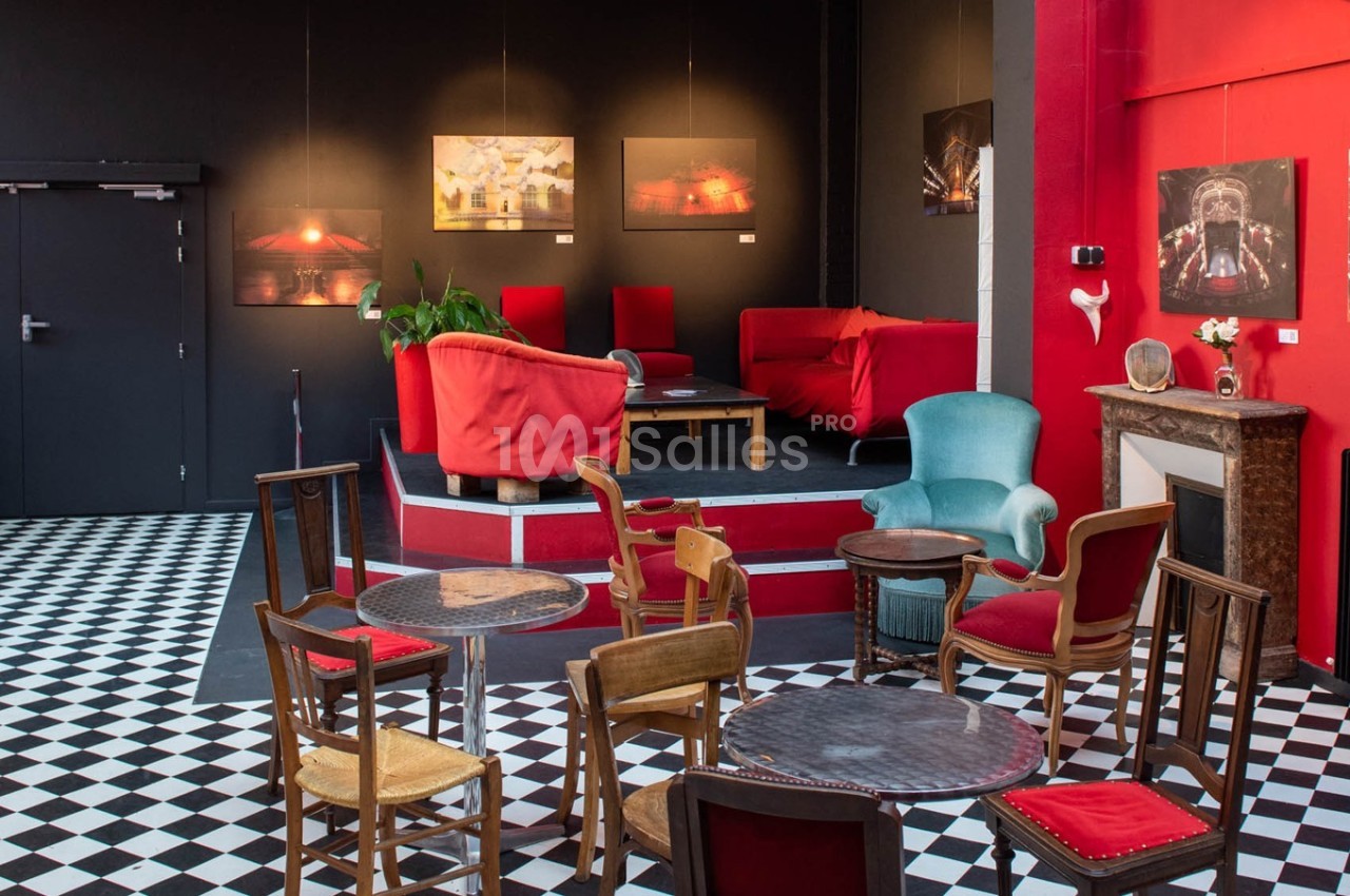Salle avec tables et chaises en bois, fauteuils rouges et verts, sol en damier noir et blanc, et tableaux accrochés au mur.