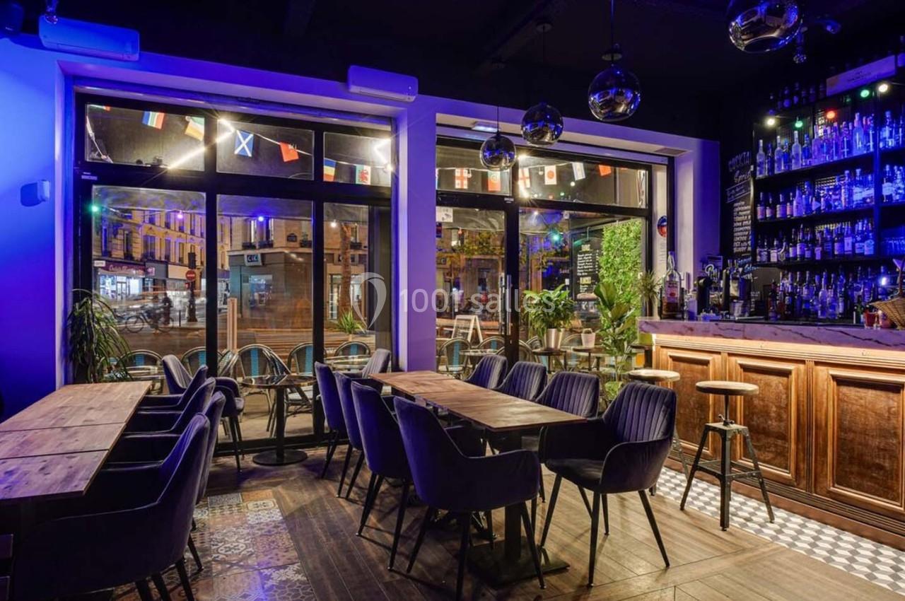 Intérieur d'un bar avec tables en bois, chaises noires, comptoir éclairé et décorations lumineuses colorées.