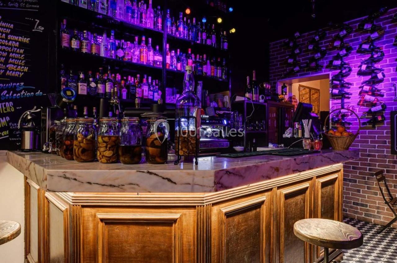 Bar avec comptoir en marbre, étagères remplies de bouteilles, éclairage violet et ambiance chaleureuse.