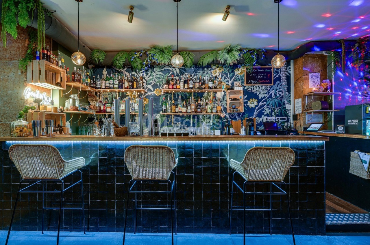 Bar avec comptoir carrelé noir, tabourets en rotin, étagères de bouteilles, décor tropical et éclairage tamisé.