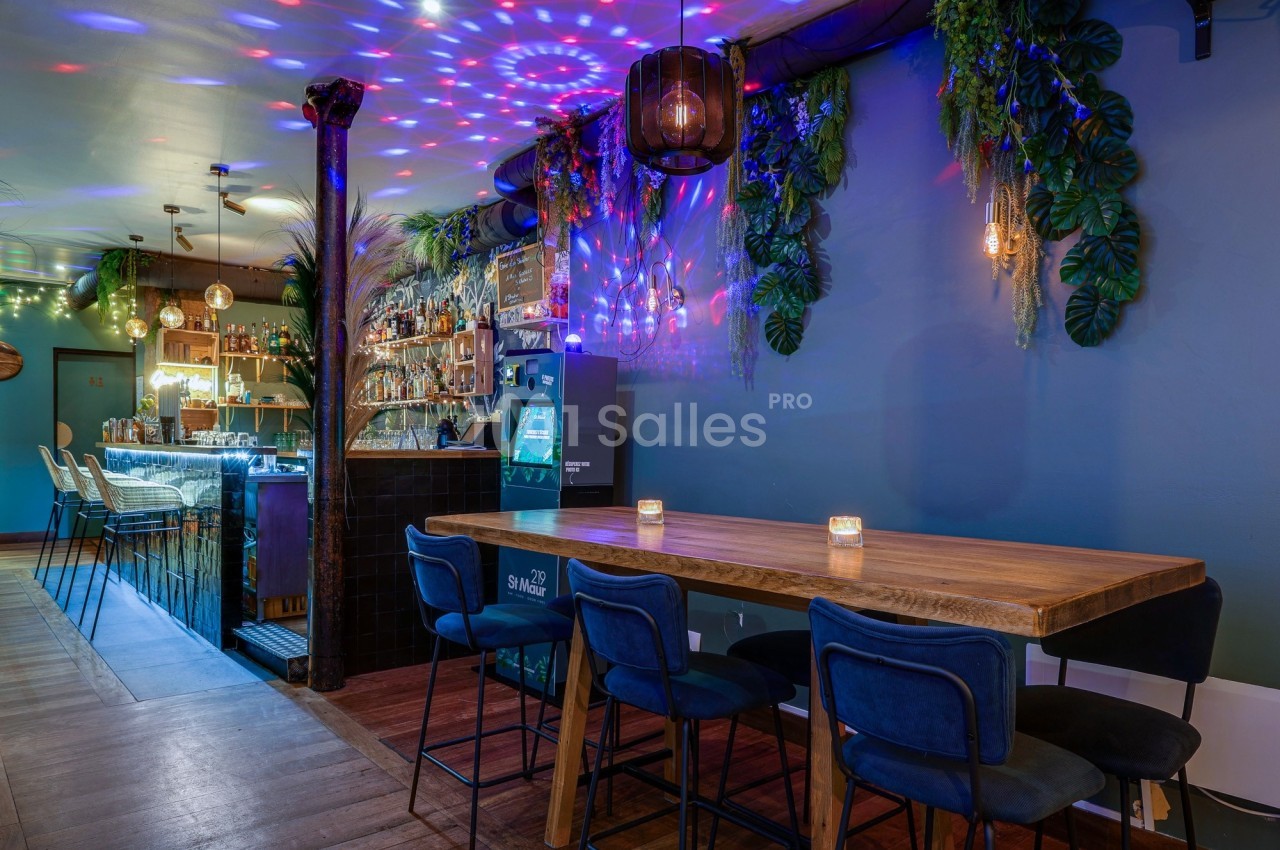Salle de bar moderne avec un long comptoir, table en bois, chaises bleues et éclairage coloré décoratif.