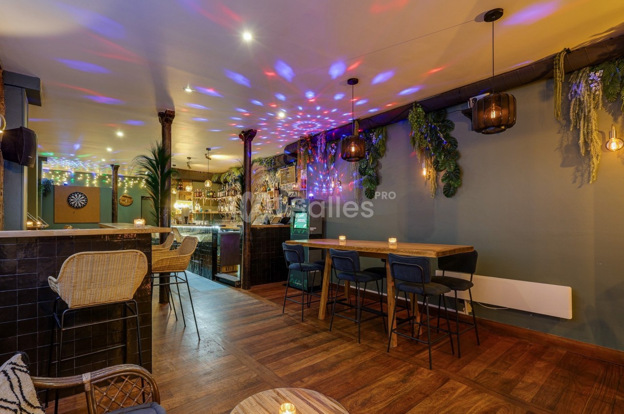 Intérieur d'un bar avec éclairage tamisé, décorations végétales, tables en bois et lumières colorées au plafond.