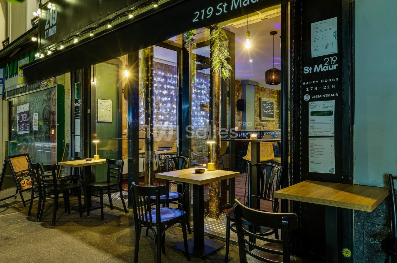 Façade d'un café-restaurant avec tables en terrasse, éclairage chaleureux et intérieur visible à travers les vitrines.