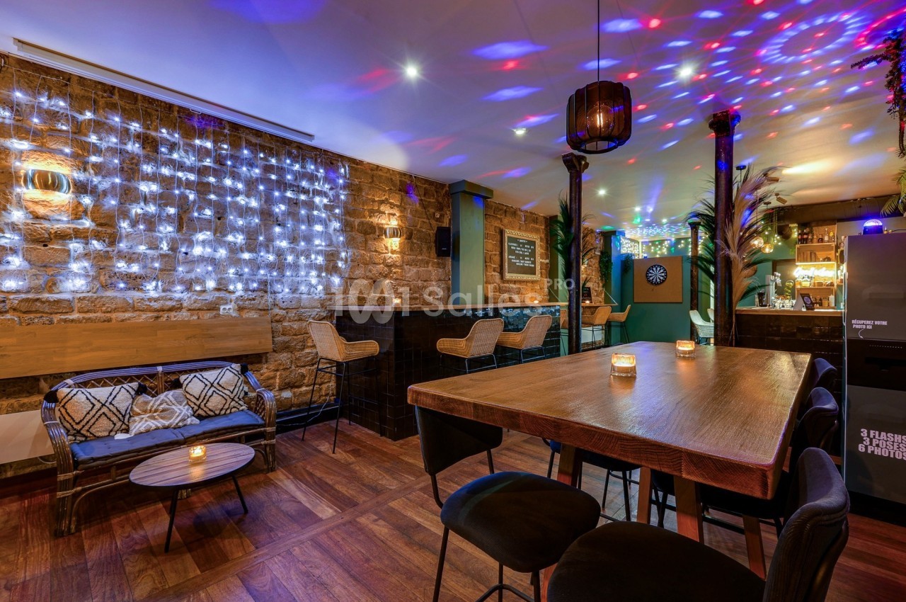Salle de bar chaleureuse avec murs en pierre, guirlandes lumineuses, table haute en bois et éclairage coloré.