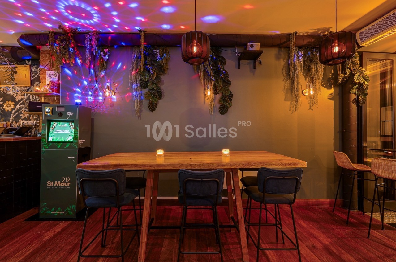Salle avec une grande table en bois, chaises hautes, éclairage tamisé, plantes suspendues et lumières colorées.