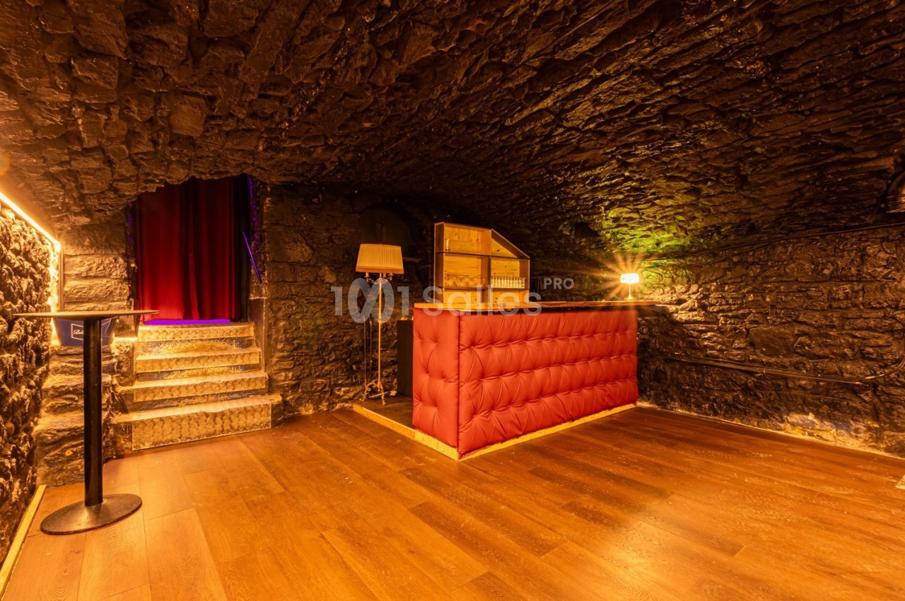 Salle voûtée en pierre avec un comptoir rouge, éclairage chaleureux et escalier menant à un rideau rouge.