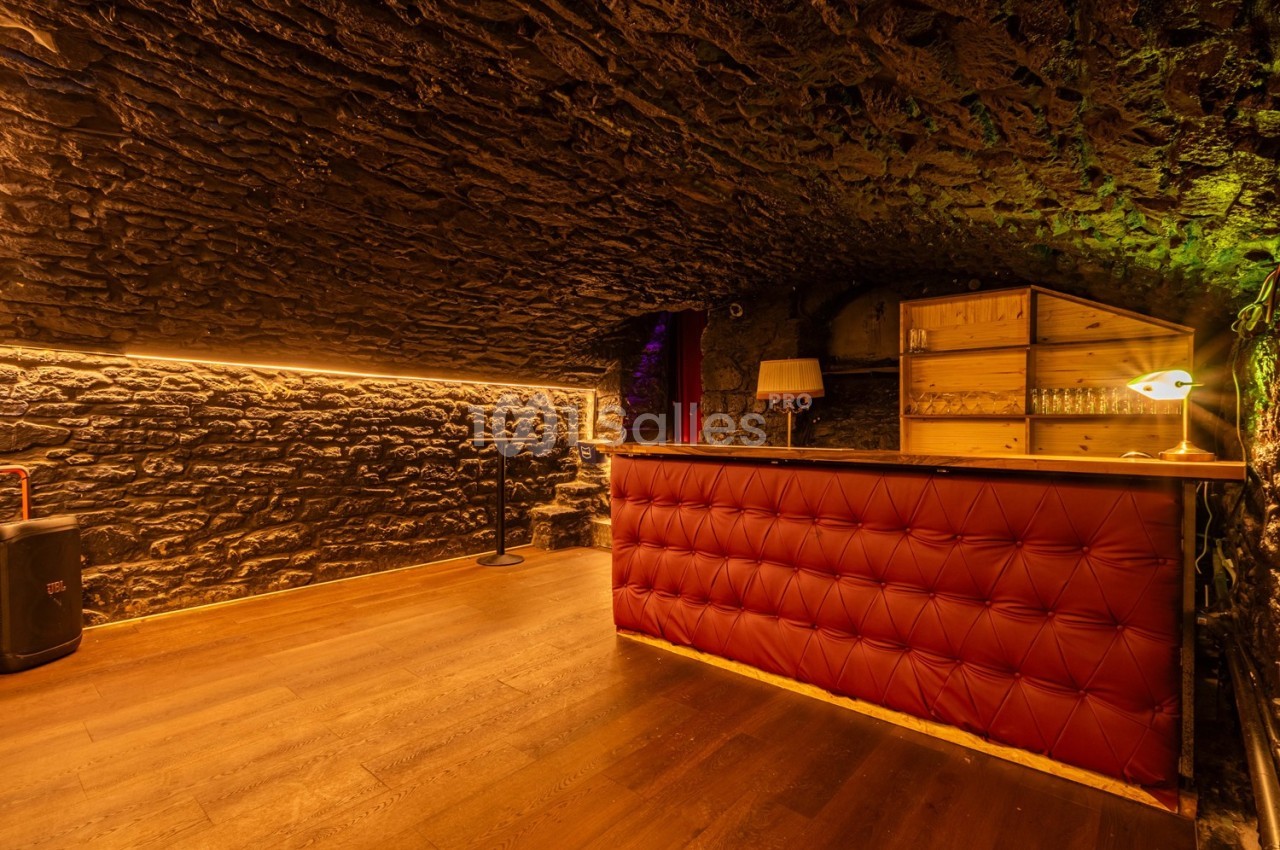Cave voûtée en pierre avec un bar en cuir rouge, éclairage chaleureux et étagères en bois à l'arrière.