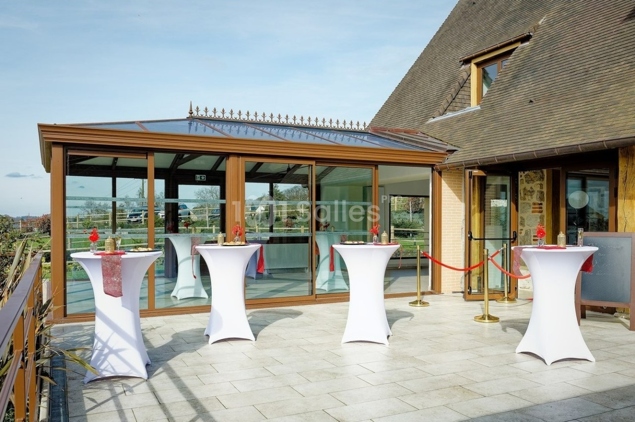 Terrasse avec tables hautes habillées de nappes blanches, devant une véranda vitrée et un bâtiment en bois.
