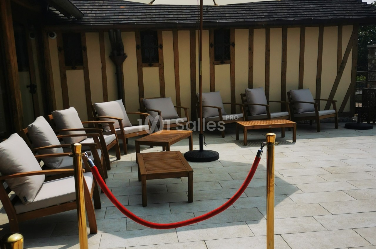 Espace extérieur avec fauteuils et tables en bois, délimité par une corde rouge et poteaux dorés.
