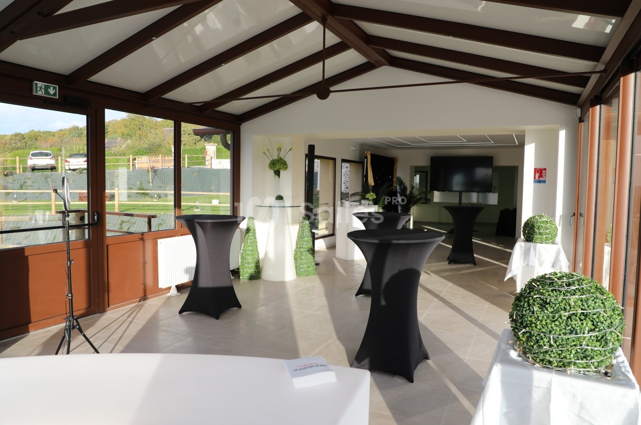 Salle lumineuse avec tables hautes noires, décorations végétales et grandes baies vitrées donnant sur l'extérieur.