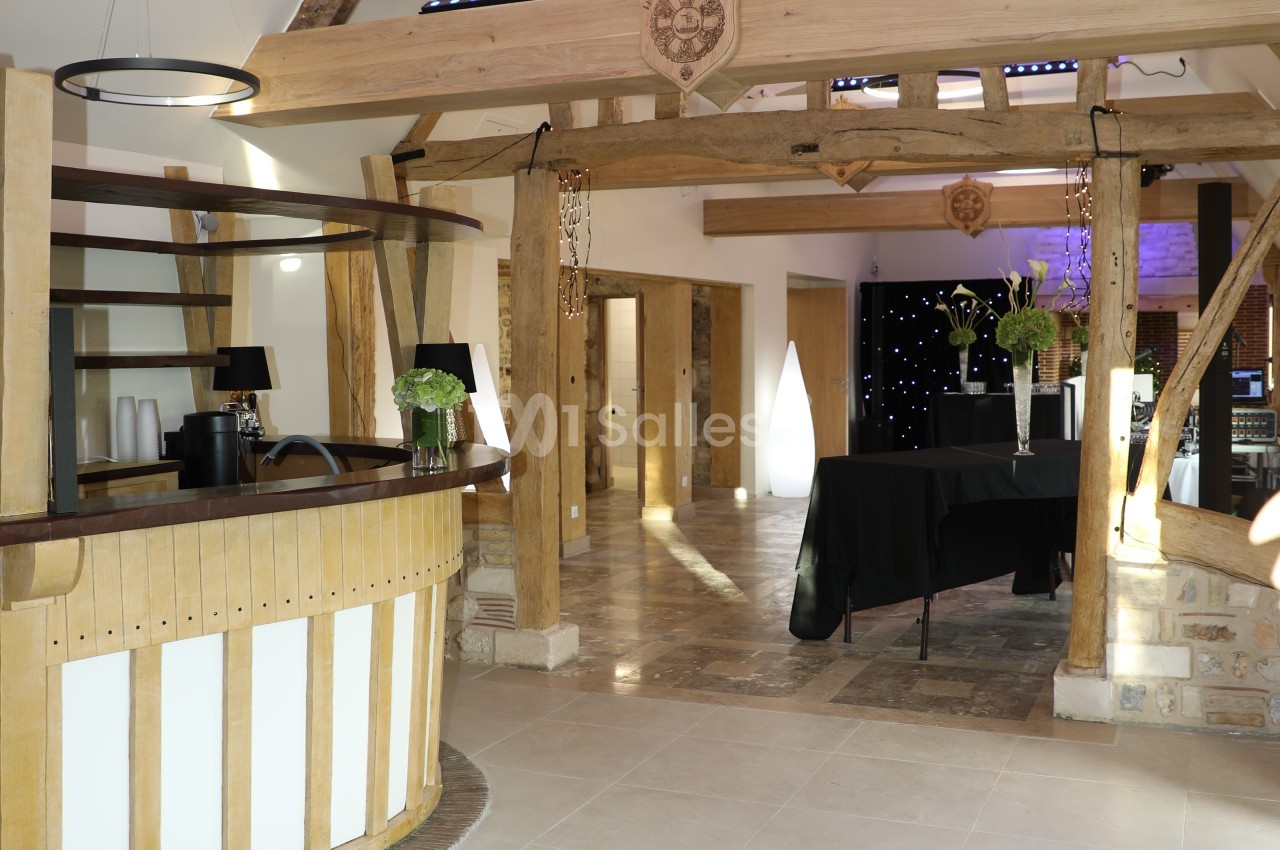 Intérieur d'une salle de réception avec poutres apparentes, comptoir en bois et tables décorées de fleurs.