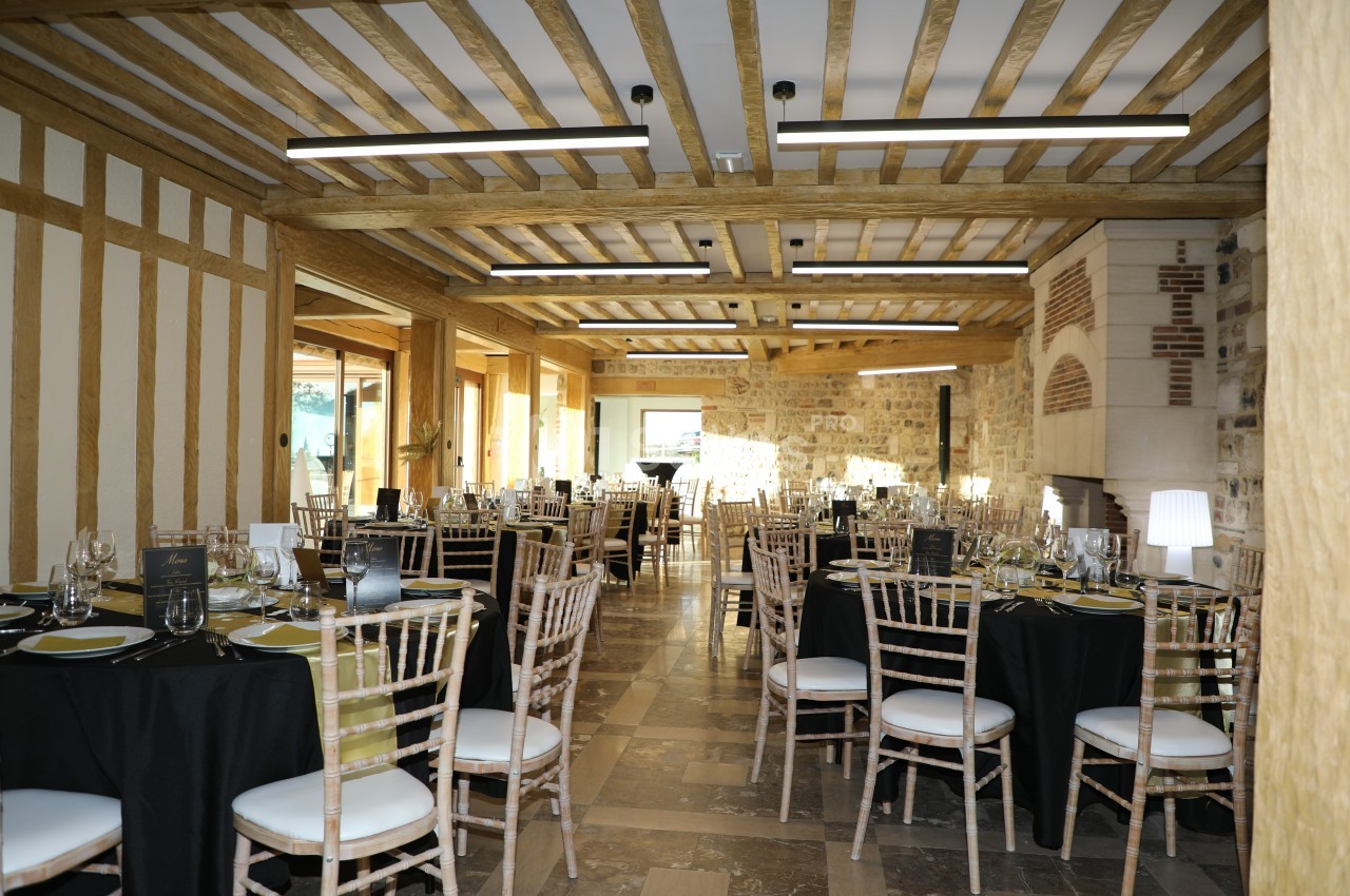 Salle de réception lumineuse avec tables dressées, chaises en bois clair et poutres apparentes au plafond.