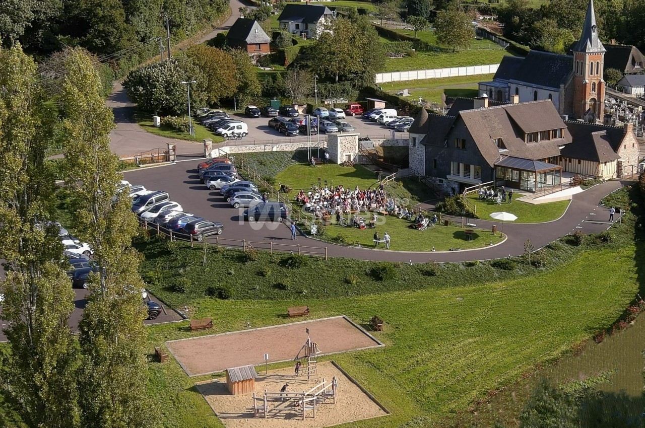 Vue aérienne d'un village avec une église, un parking, une aire de jeux et un rassemblement de personnes en extérieur.