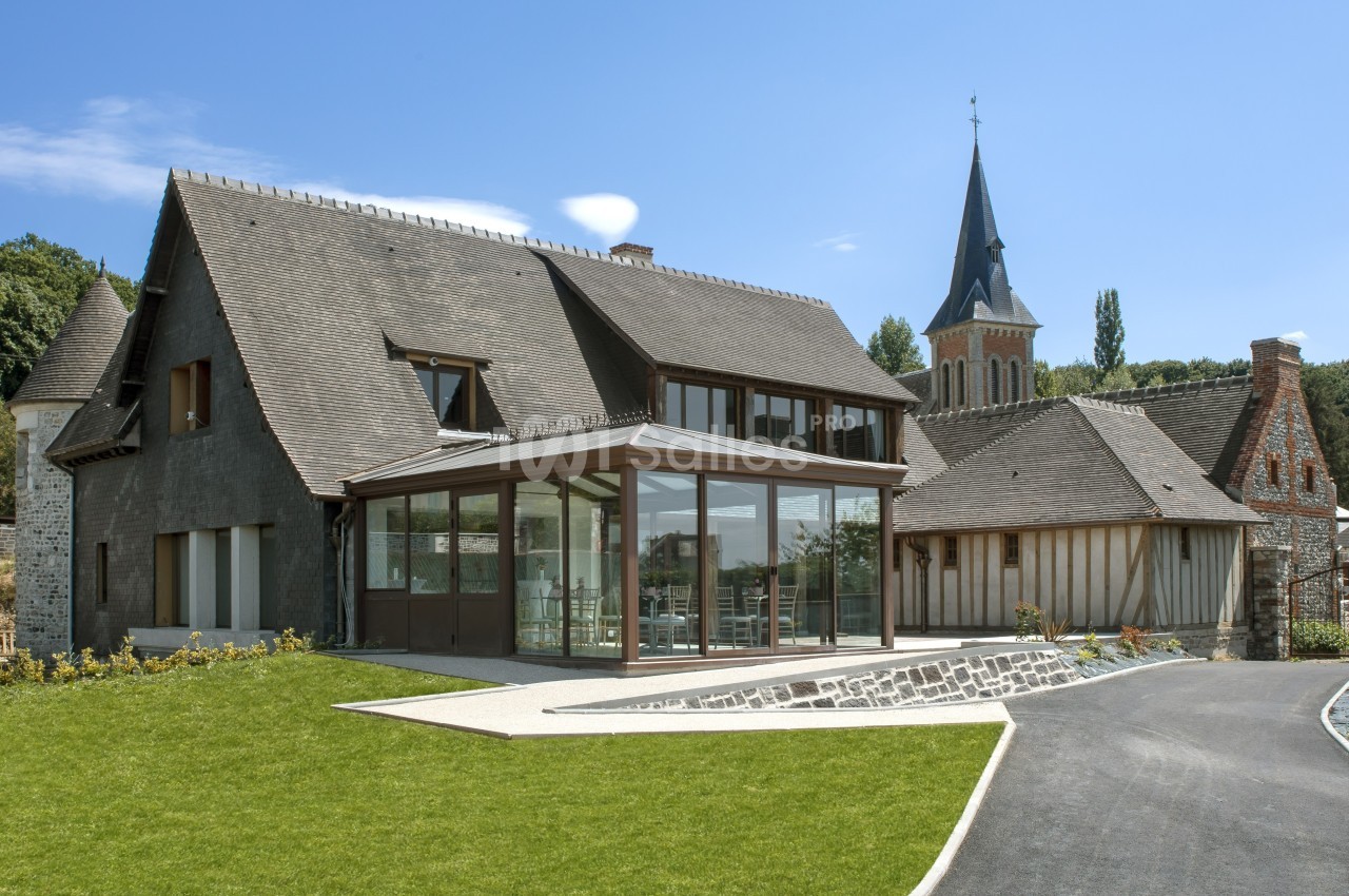 Grande maison moderne avec façade en pierre, extension vitrée et jardin, située près d'une église avec clocher.