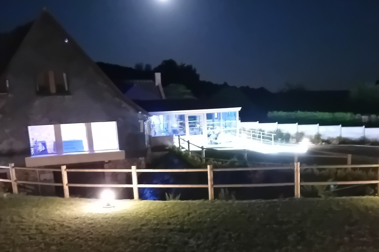 Maison avec extension moderne éclairée la nuit, entourée d'une clôture en bois et d'un jardin.
