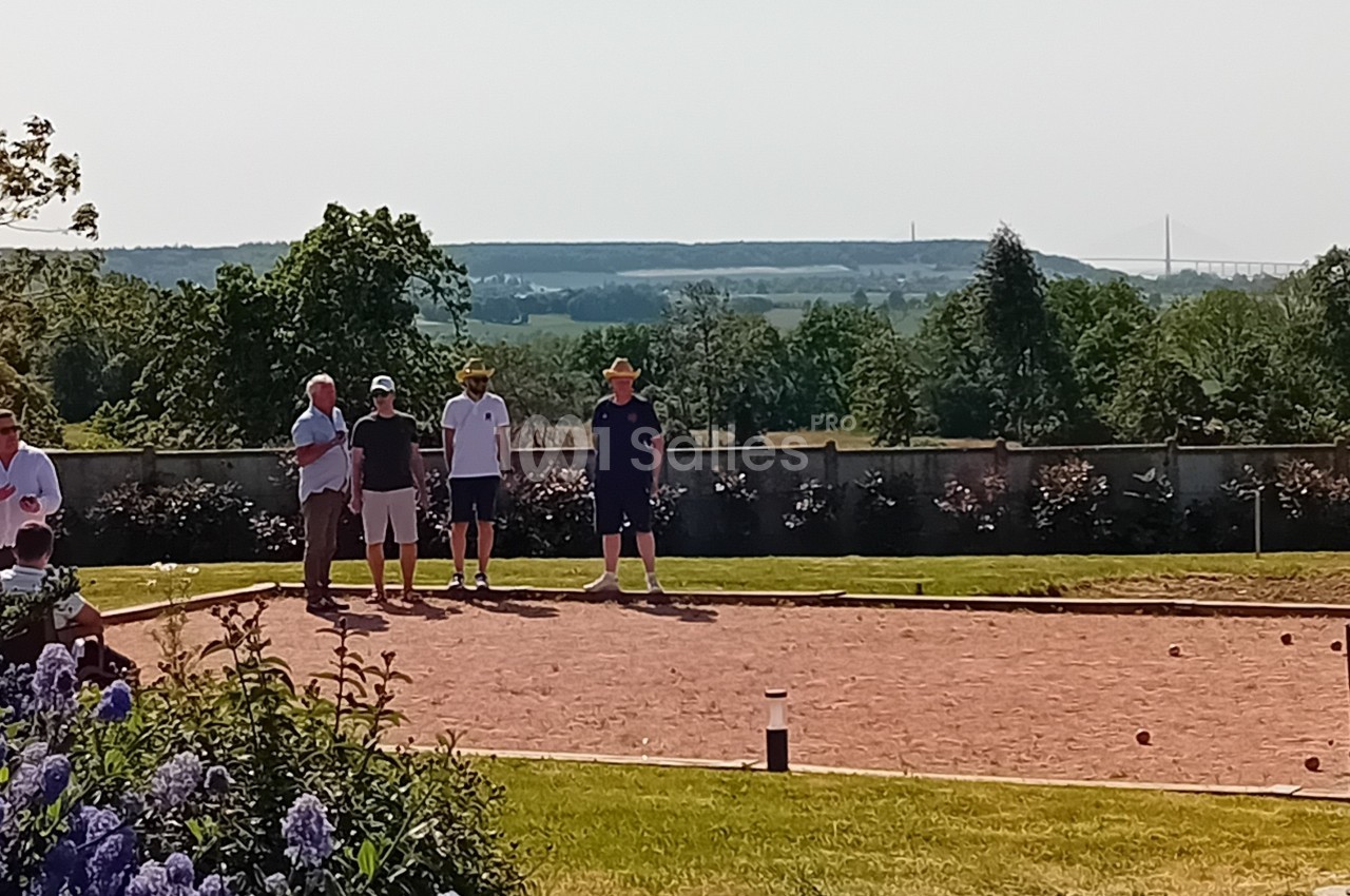 Des personnes jouent à la pétanque sur un terrain ensoleillé, avec un paysage verdoyant et un pont visible au loin.