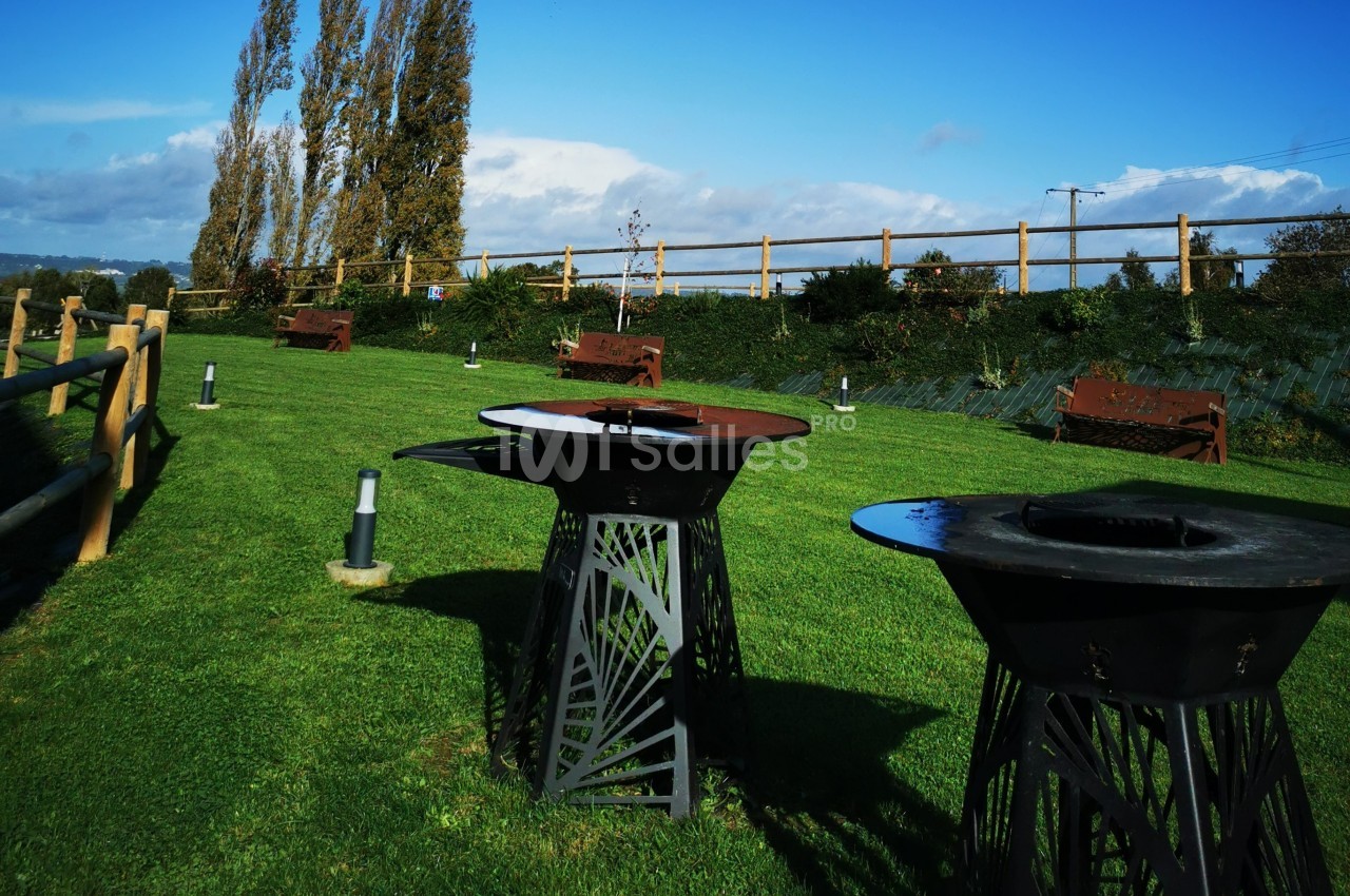 Barbecues en métal sur une pelouse verte, entourés de bancs en bois et d'une clôture en bois sous un ciel dégagé.