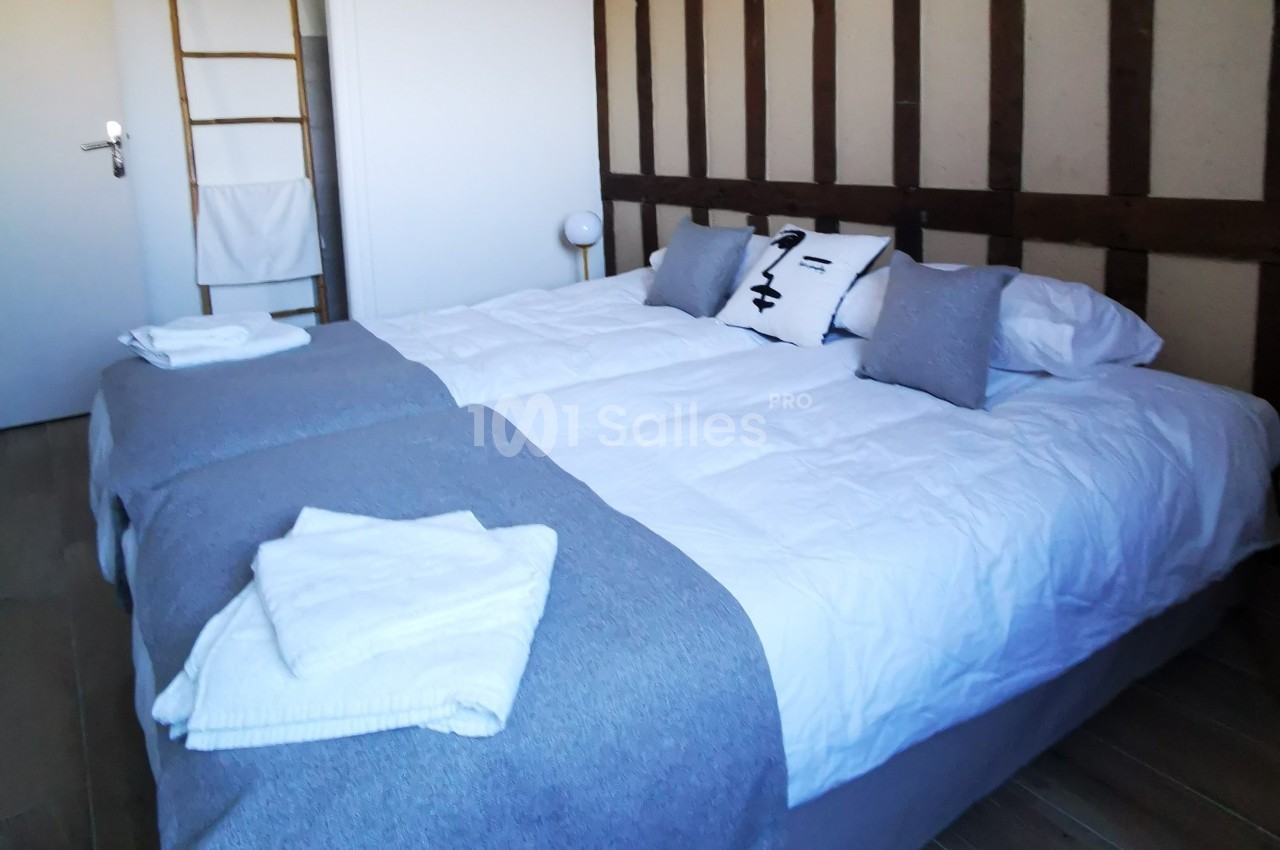 Lit double avec draps blancs, couvertures grises, oreillers assortis et serviettes pliées dans une chambre lumineuse.