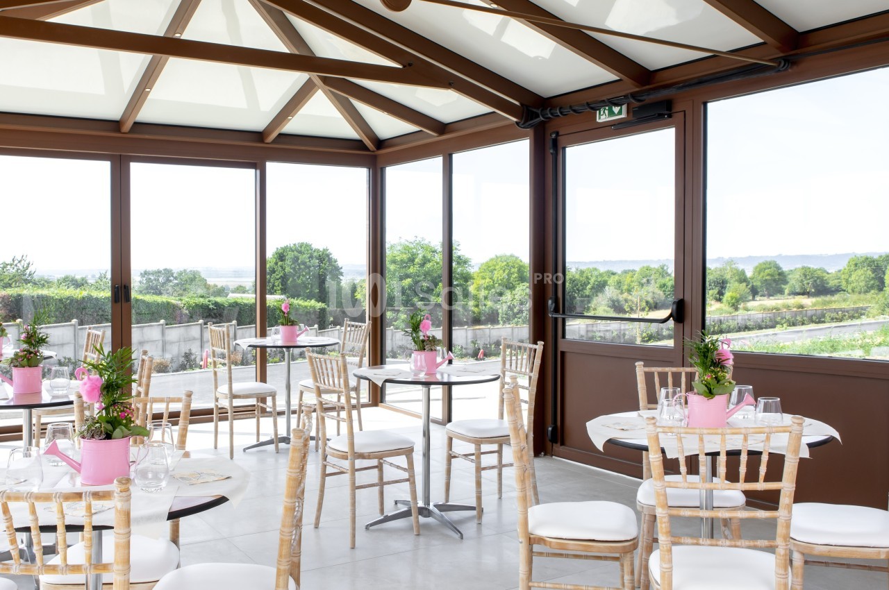 Salle lumineuse avec baies vitrées, tables rondes décorées de fleurs roses et chaises en bois, vue sur un paysage verdoyant.