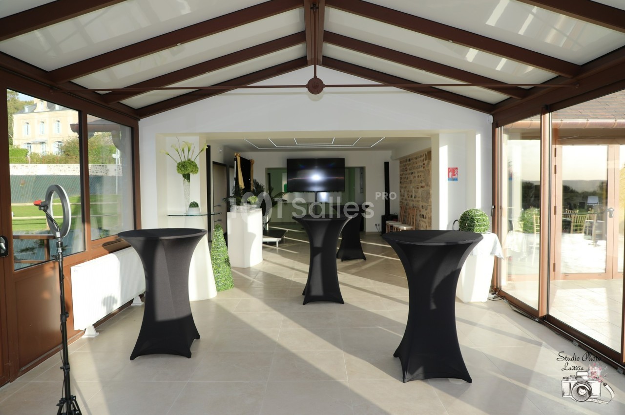 Salle lumineuse avec baies vitrées, tables hautes noires et écran en fond, décorée de fleurs et ouverte sur un jardin.