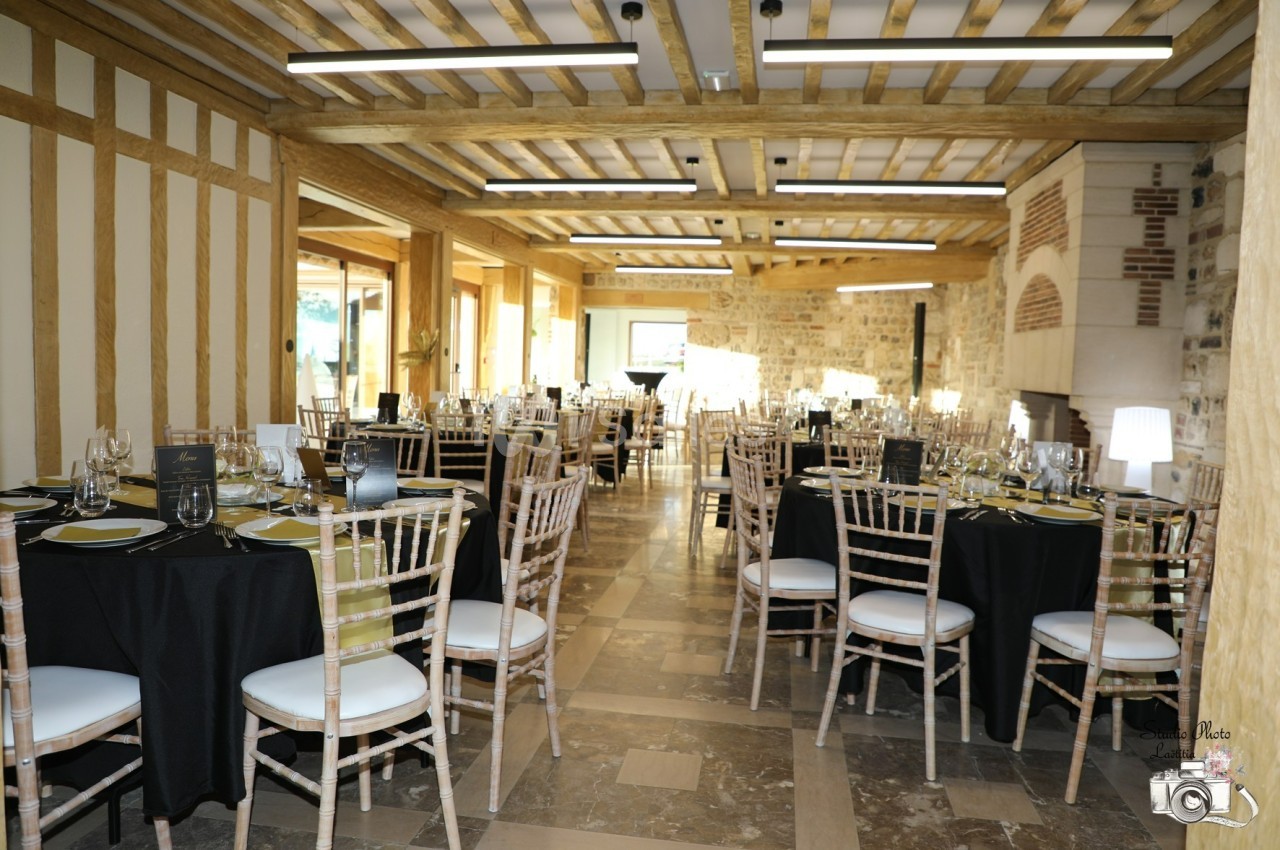 Salle de réception lumineuse avec tables dressées, nappes noires et chaises en bois, dans un décor rustique en pierre et…