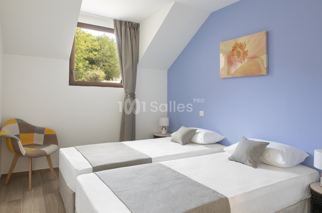 Deux lits simples avec draps blancs et couvre-lits gris dans une chambre lumineuse aux murs bleus et blancs.