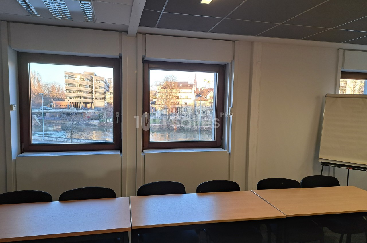 Salle de réunion avec des tables et des chaises alignées, vue sur des bâtiments et un cours d'eau à travers les fenêtres.