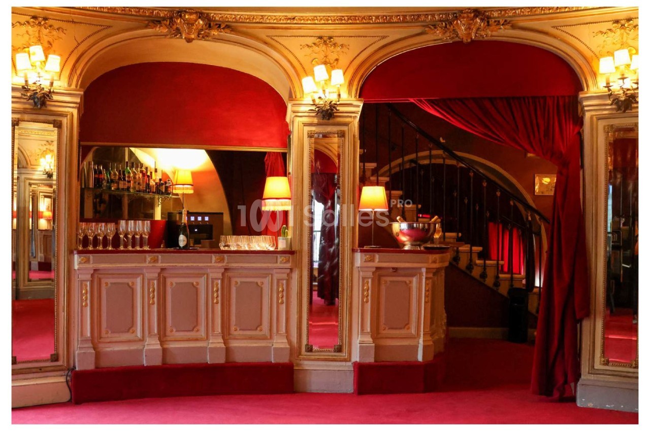 Bar élégant avec comptoir en bois sculpté, miroirs, lumières tamisées et rideaux rouges dans un décor classique.