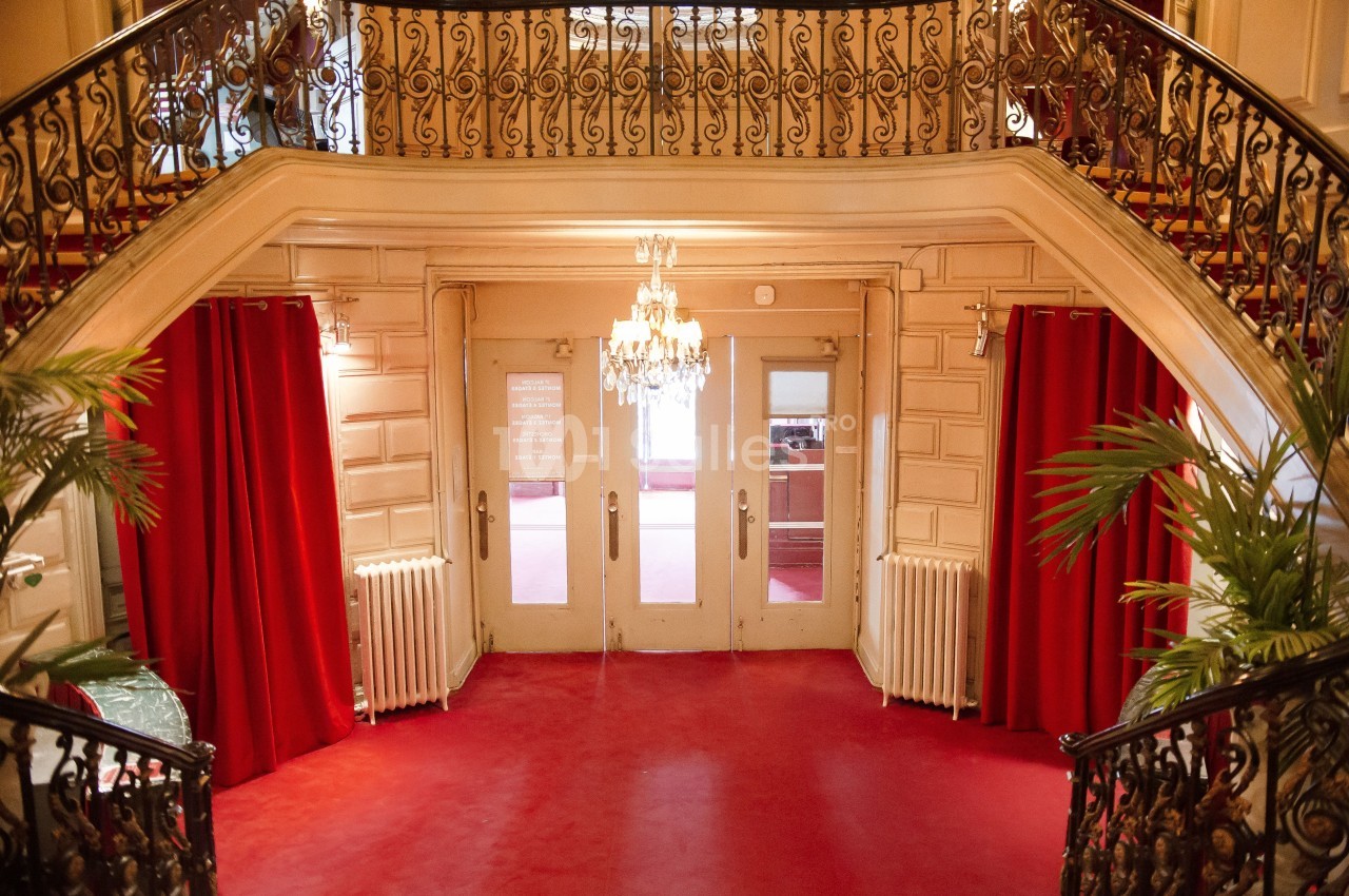 Hall d'entrée élégant avec tapis rouge, escalier en fer forgé, rideaux rouges et lustre central.