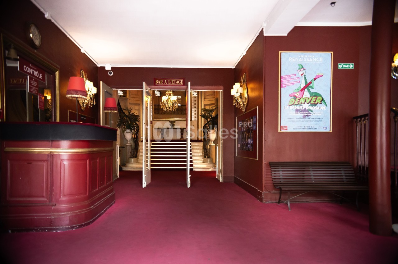 Entrée d'un théâtre avec comptoir rouge, escalier central, affiches murales et éclairage tamisé.