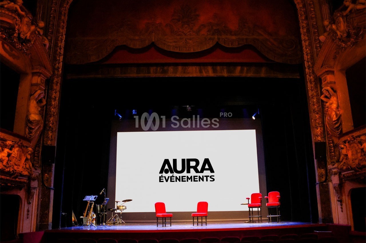 Scène de théâtre avec écran affichant ’Aura Événements’, quatre fauteuils rouges et des instruments de musique.