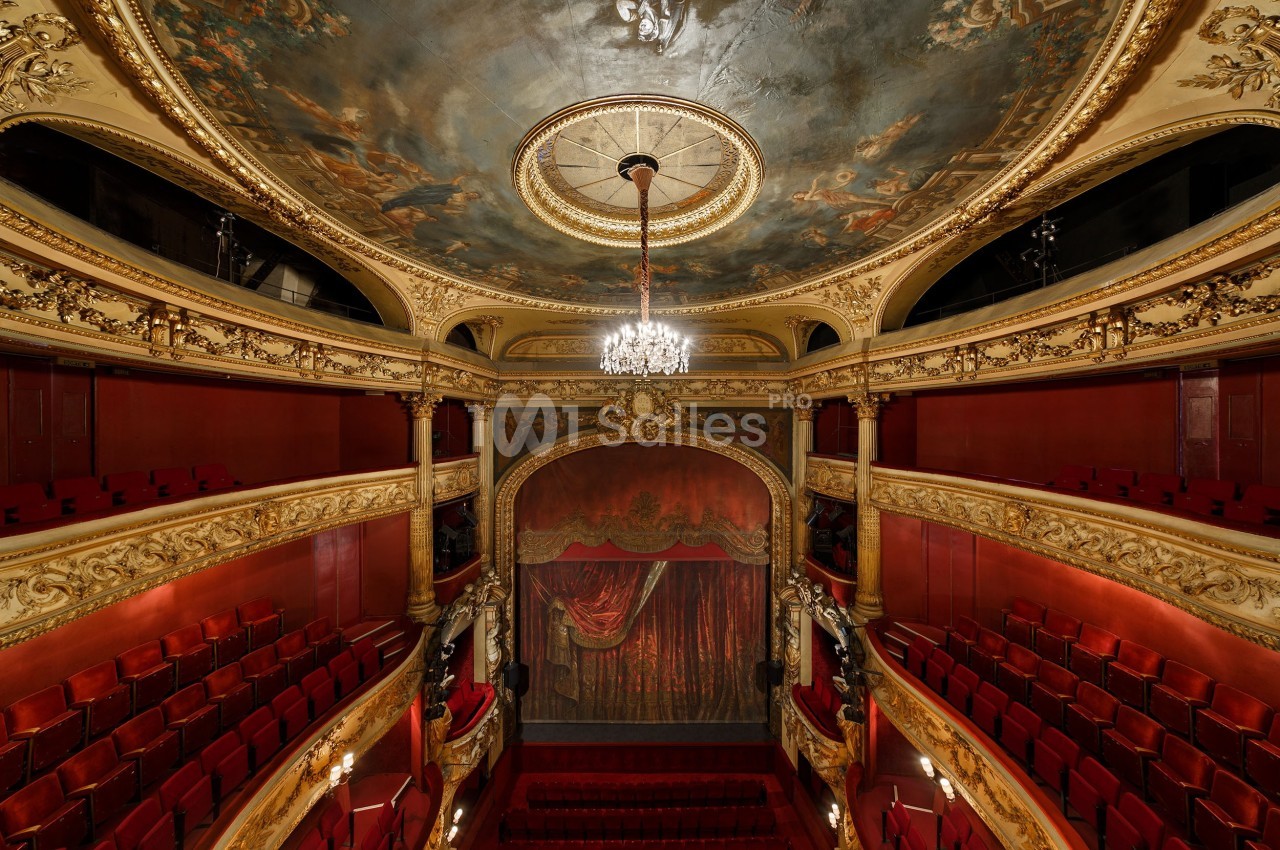 Intérieur d'un théâtre ancien avec balcon, plafond peint, lustre central et rideau de scène rouge.