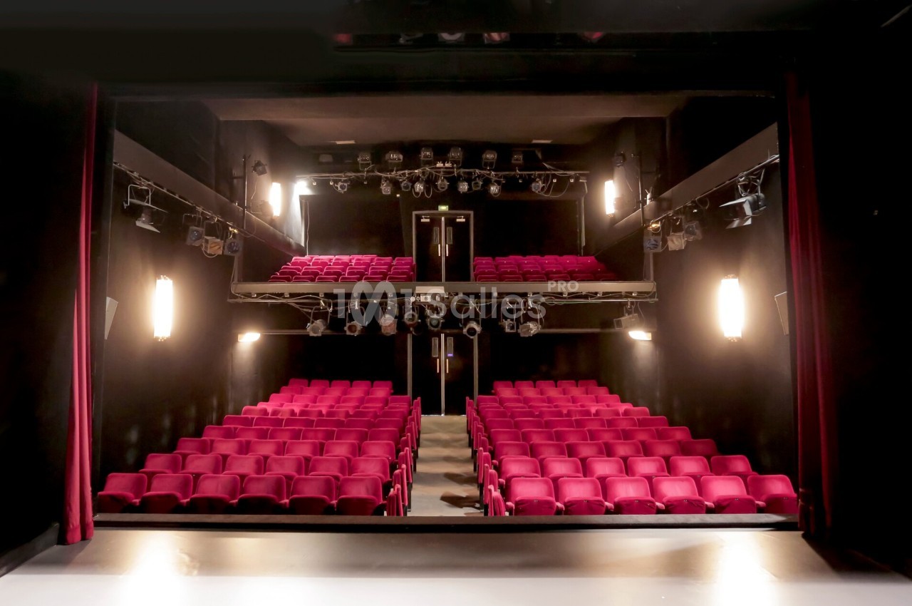 Salle de théâtre avec rangées de sièges rouges, scène au premier plan et éclairages suspendus.