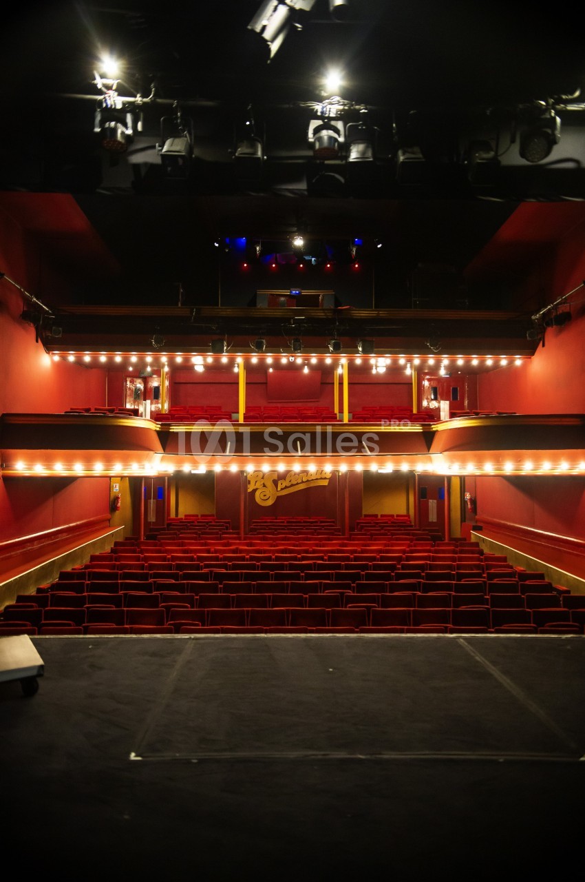 Vue depuis la scène d'un théâtre vide avec des rangées de sièges rouges et des balcons éclairés.