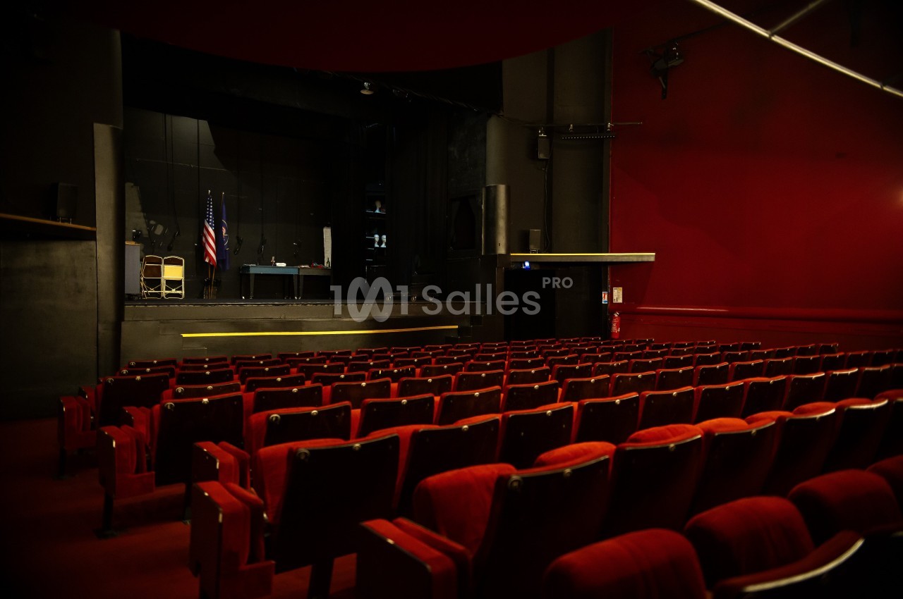 Salle de théâtre vide avec des rangées de sièges rouges face à une scène sombre équipée de quelques accessoires.