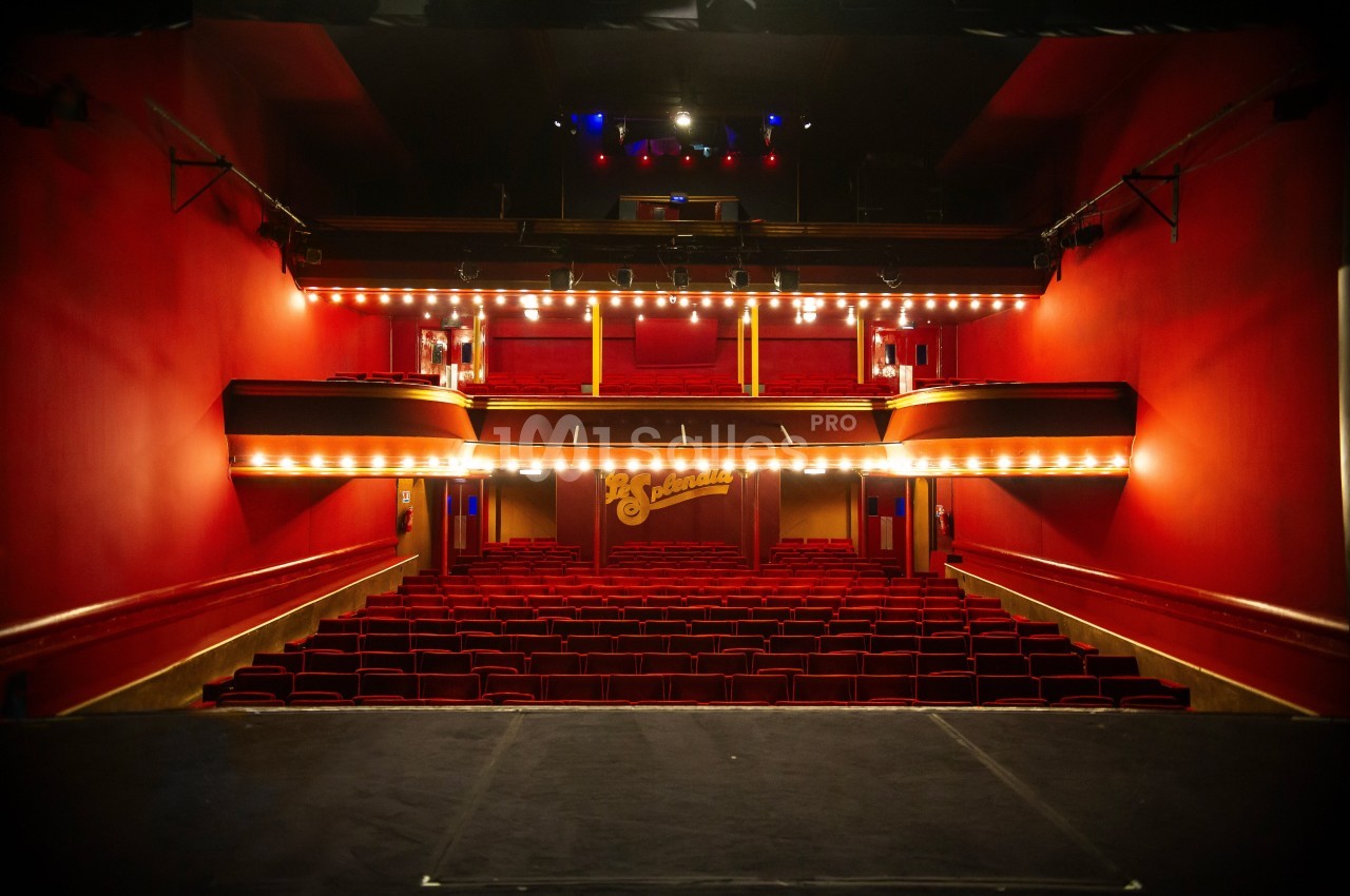 Salle de théâtre vide avec sièges rouges, balcons éclairés et scène vue depuis les coulisses.