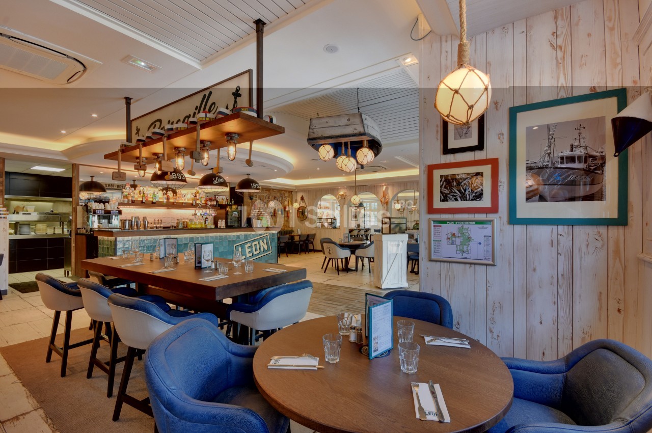 Salle de restaurant lumineuse avec tables dressées, chaises colorées et décoration murale maritime.
