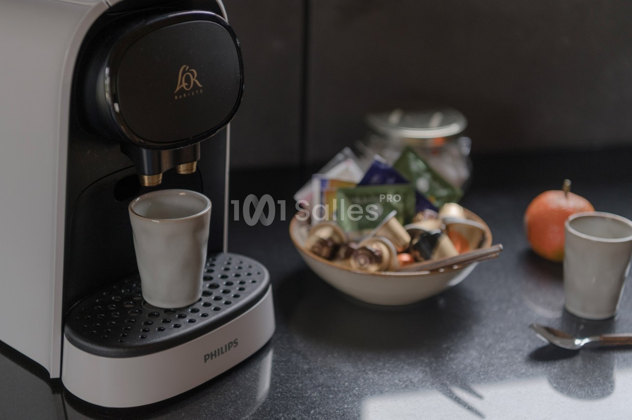 Machine à café avec une tasse en céramique, bol contenant des capsules et sachets de thé sur un plan de travail sombre.