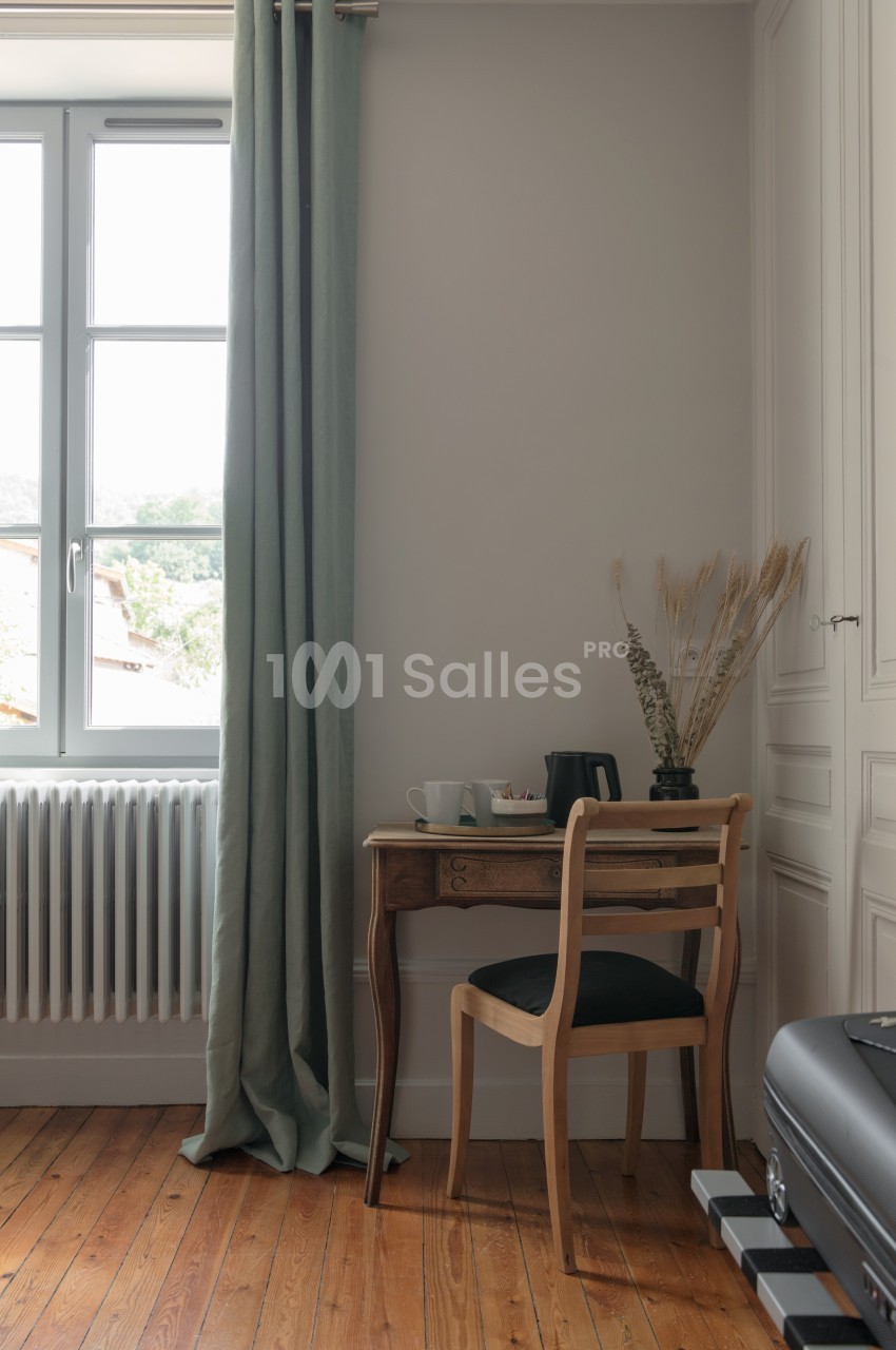 Petit bureau en bois avec une chaise, une bouilloire et une tasse, près d'une fenêtre avec rideaux verts et un radiateur.