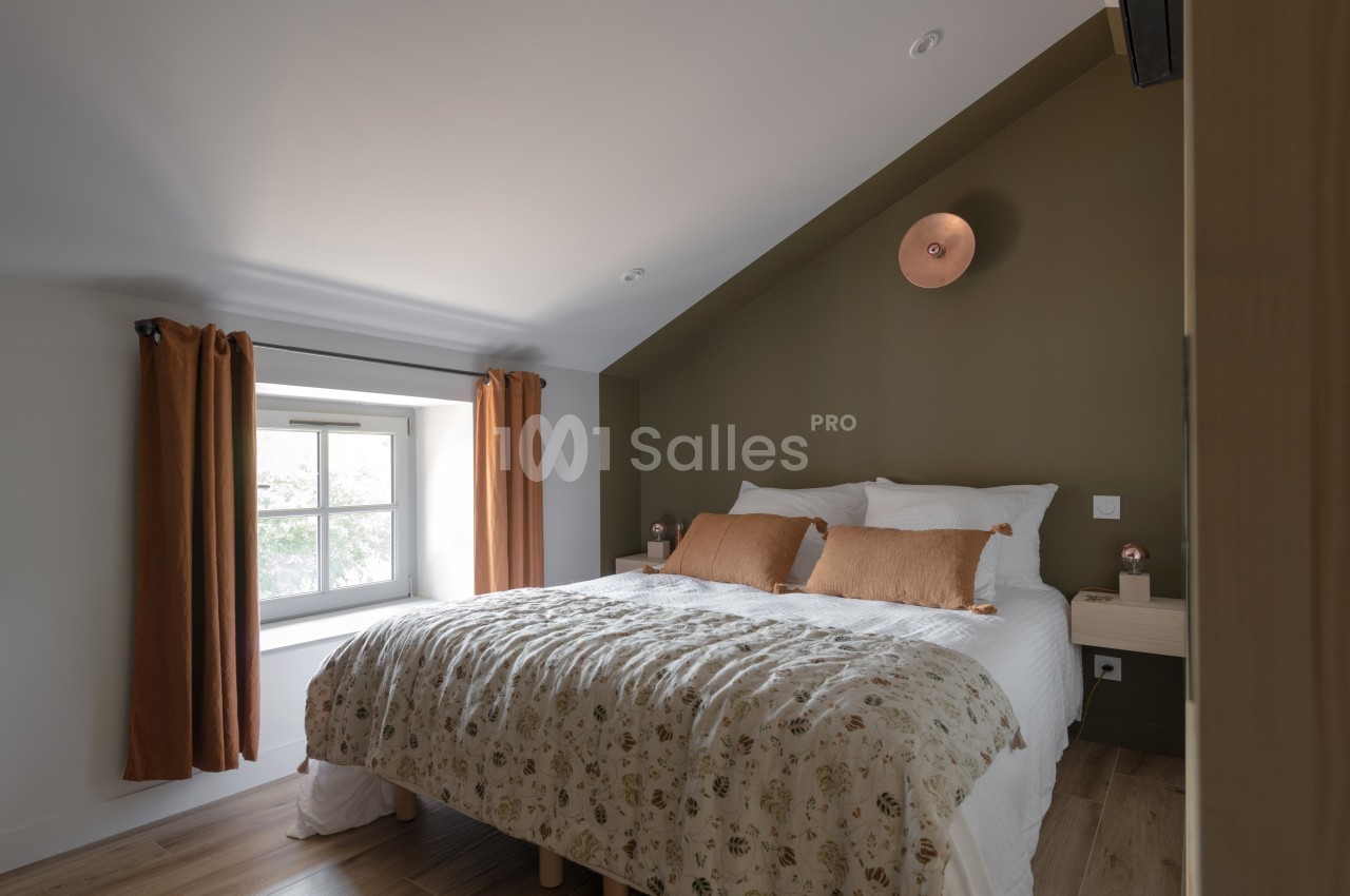 Chambre lumineuse avec lit double, mur vert olive, fenêtre avec rideaux orange et parquet au sol.