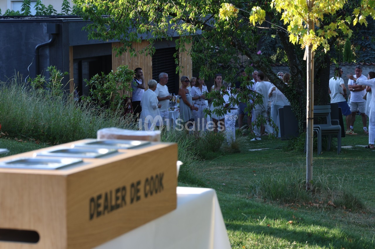 Groupe de personnes en tenue blanche rassemblées dans un jardin, avec une boîte en bois marquée ’Dealer de Cook’ au premier…