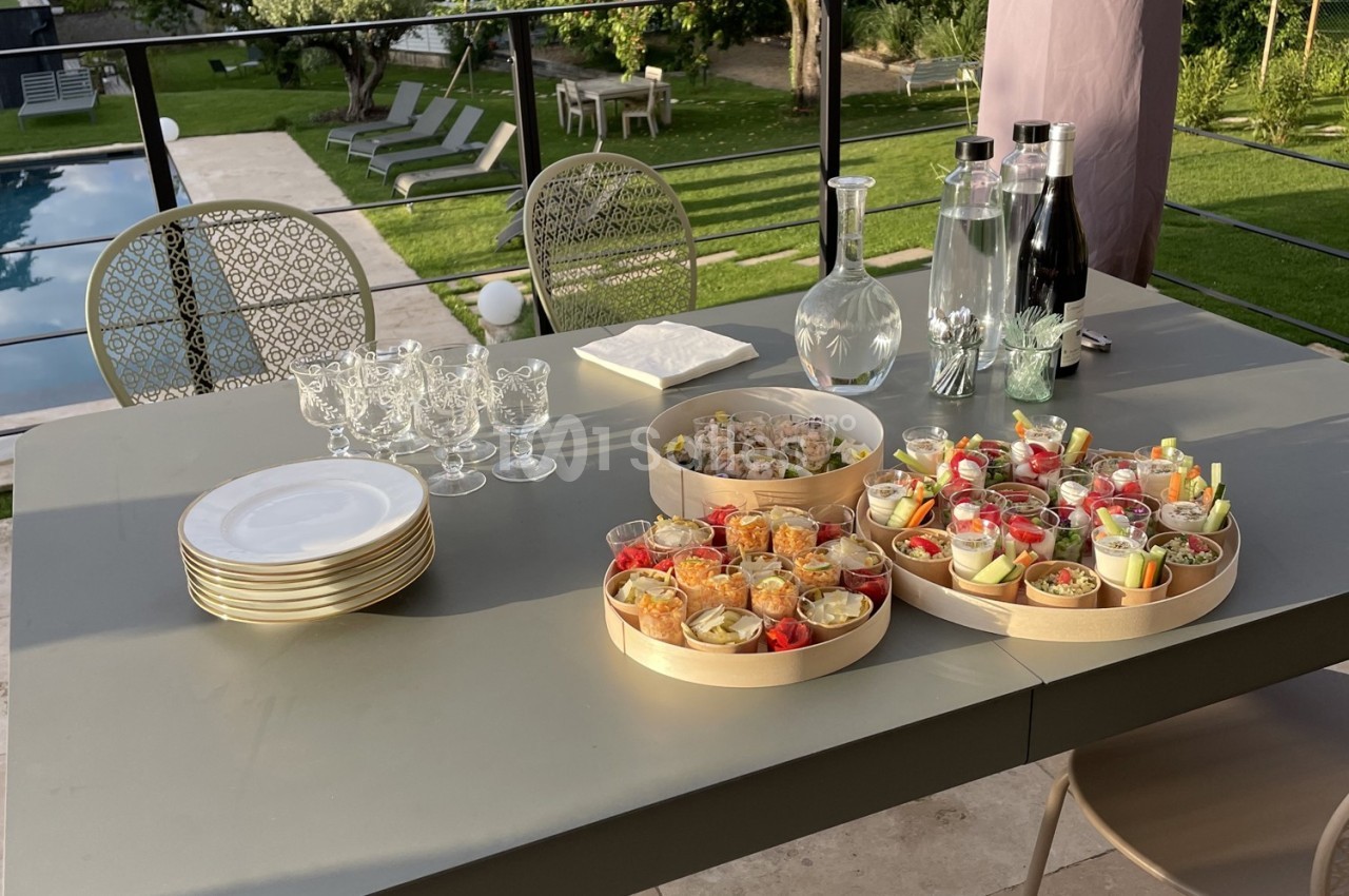 Plateau de verrines colorées et carafes sur une table dressée en extérieur, avec vue sur un jardin et une piscine.