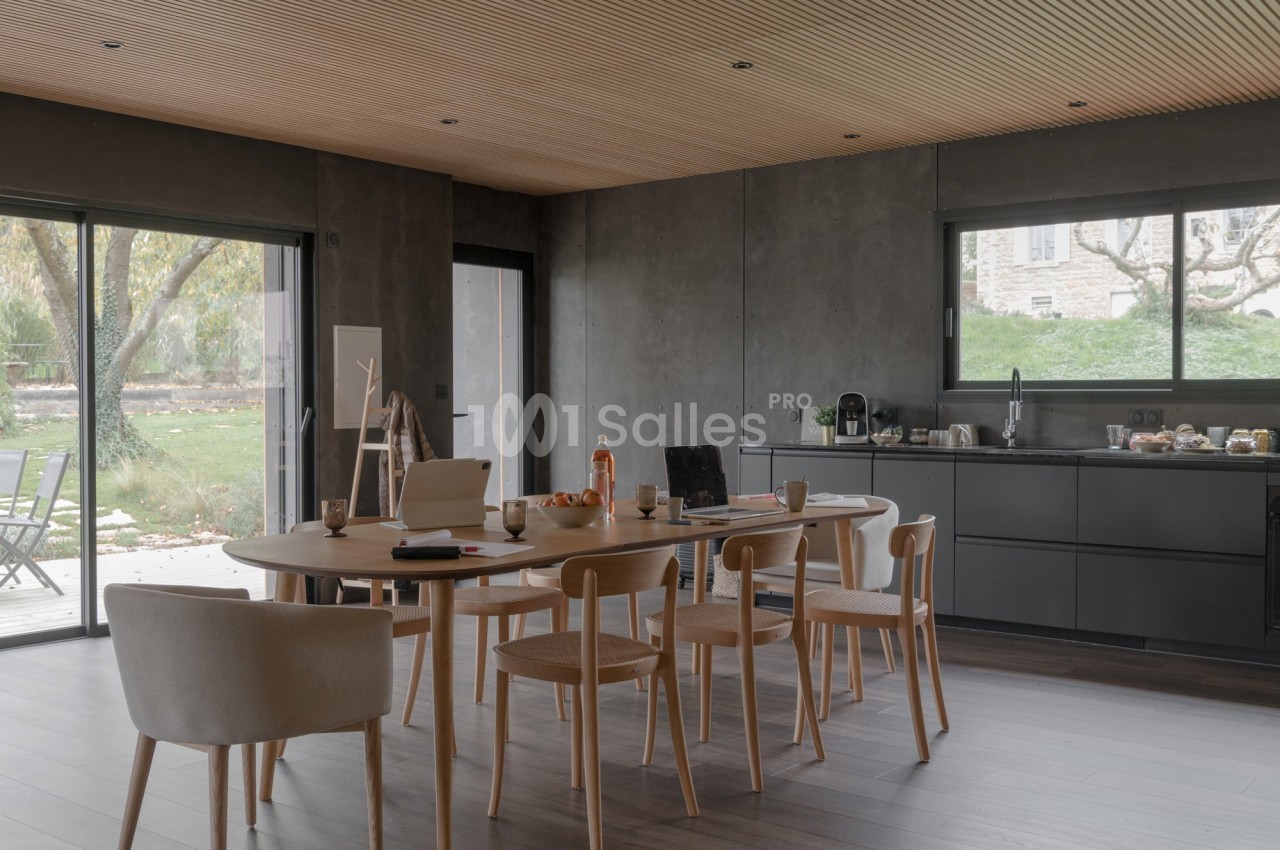 Salle à manger moderne avec table en bois, chaises assorties, cuisine ouverte et vue sur un jardin arboré.