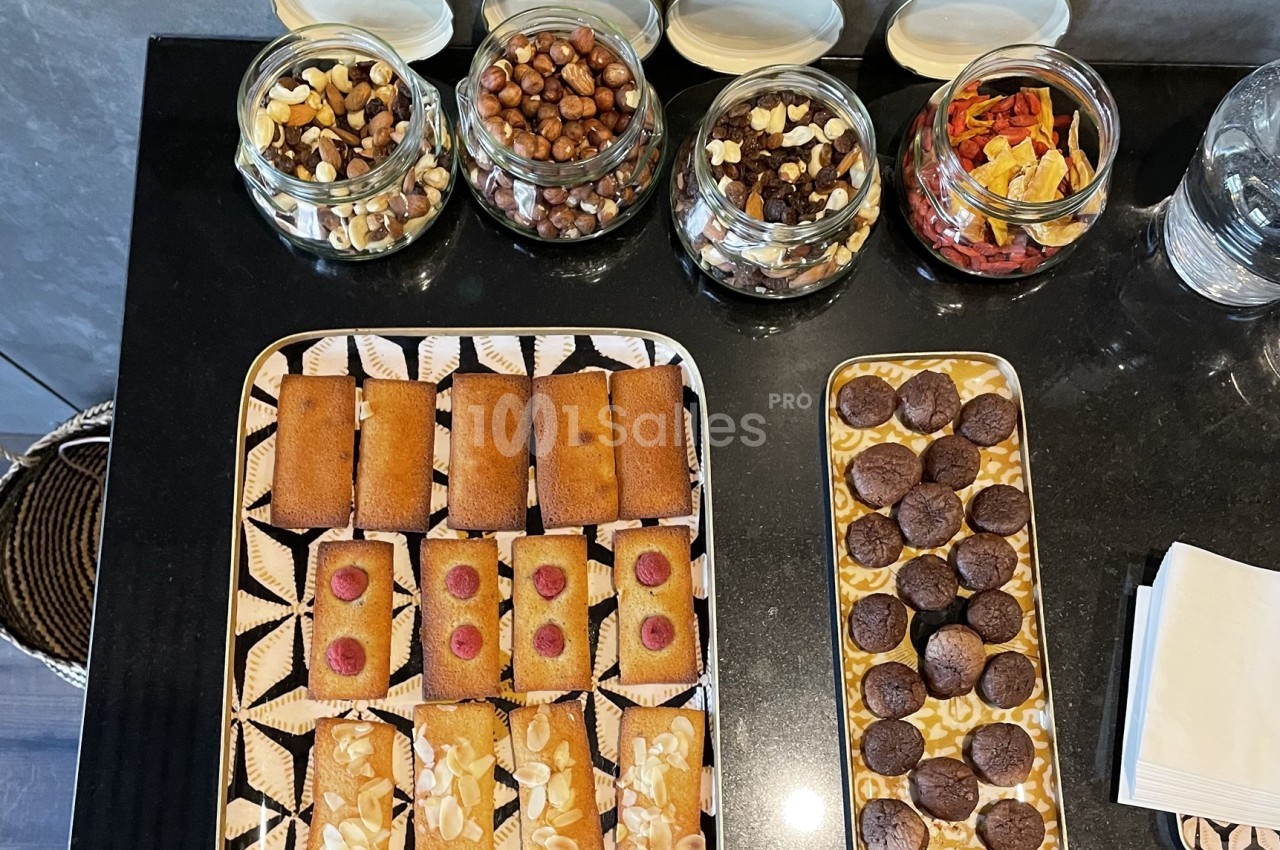 Plateaux de financiers aux amandes et framboises, biscuits au chocolat, avec pots de fruits secs en arrière-plan.