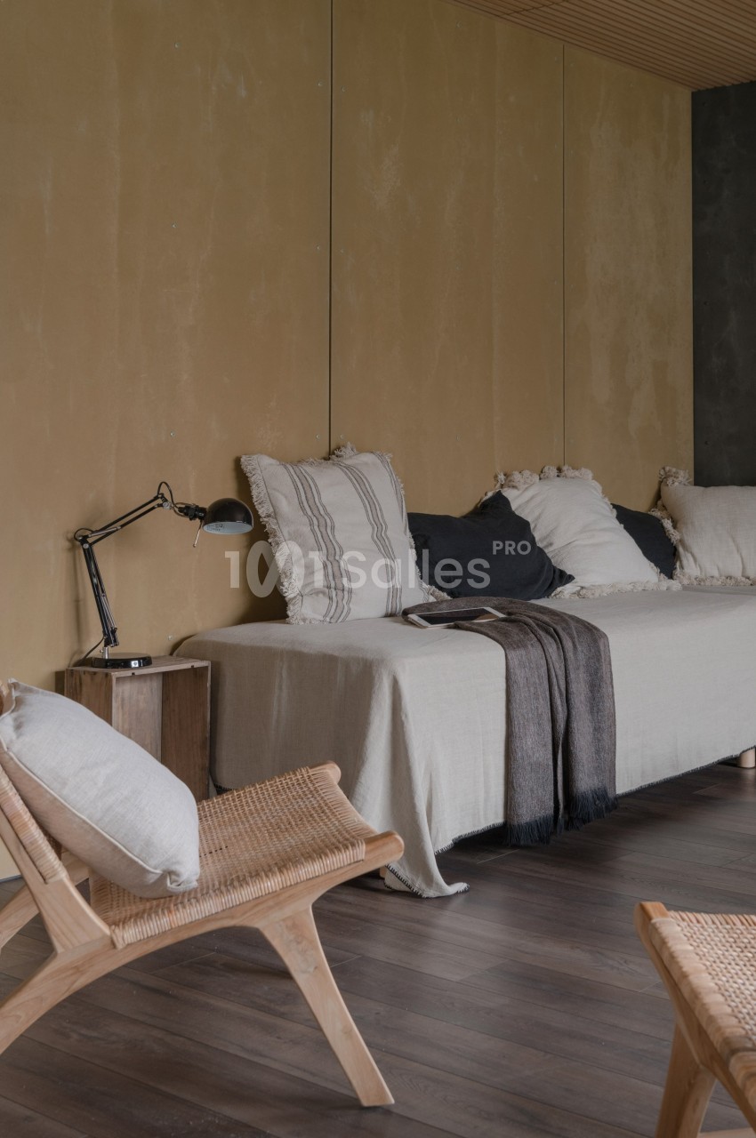 Salon minimaliste avec canapé beige, coussins texturés, plaid, lampe de lecture et fauteuil en bois sur parquet sombre.