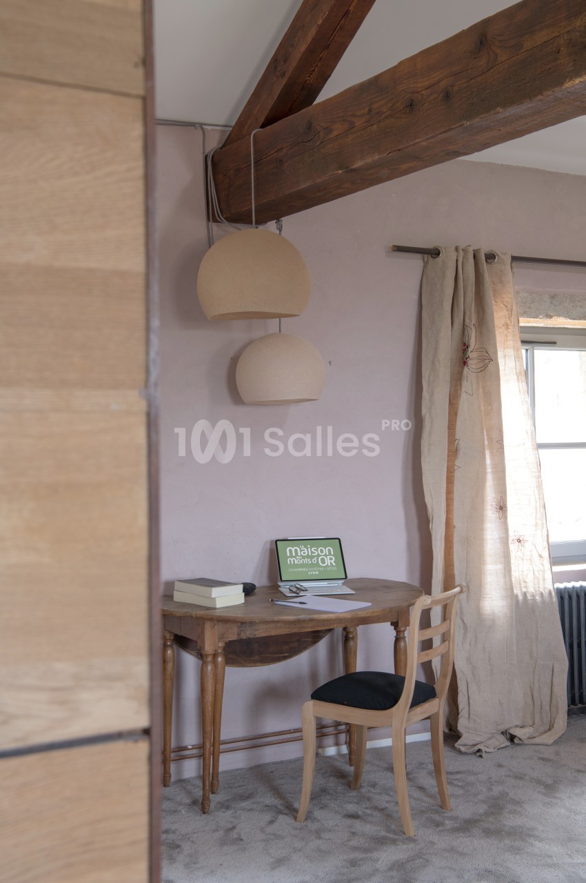 Bureau en bois avec chaise noire, lampe suspendue, rideaux en lin et ordinateur portable sur une table près d'une fenêtre.